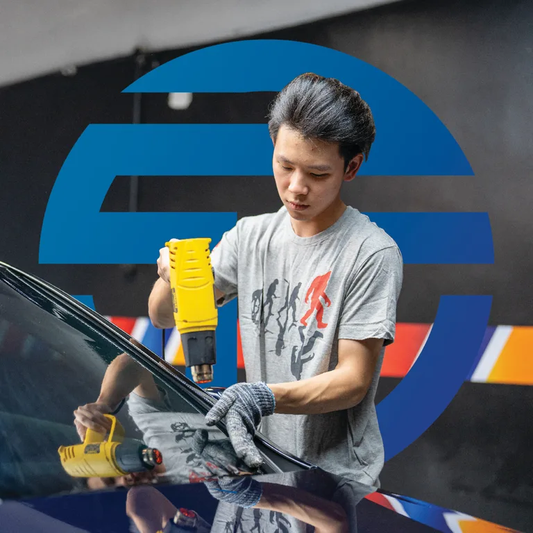 Technician using a heat gun on a car windscreen, set against TGA's oversized blue logomark